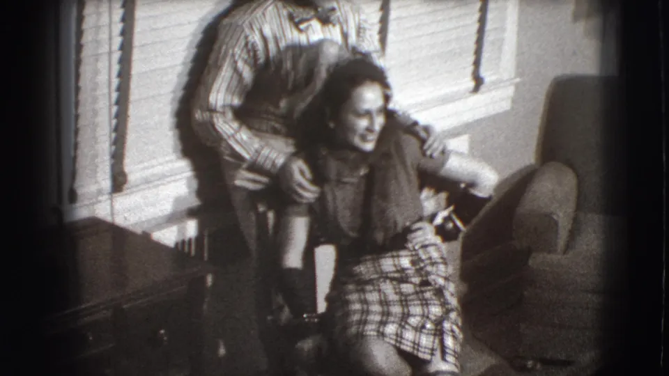 At the end of the clip, the man stands partly behind the seated woman as she laughs beside a chair in the Joliet home interior.