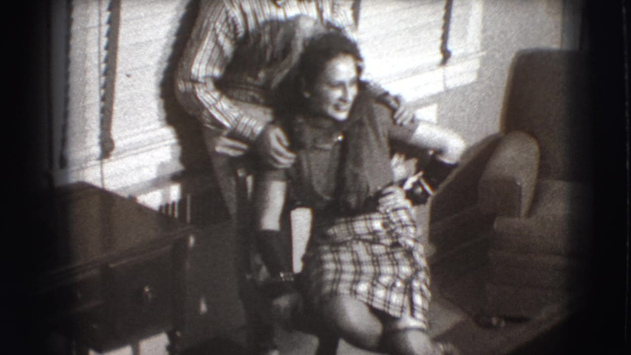 Man standing behind a laughing woman seated indoors near window blinds.