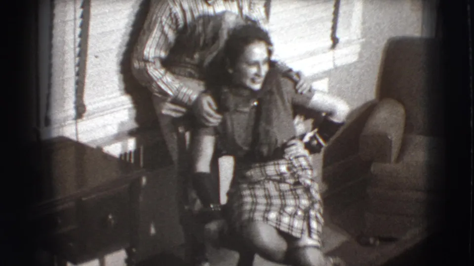 In a 1941 Joliet home-movie frame, a man leans over a seated laughing woman in a living room with blinds and an armchair.