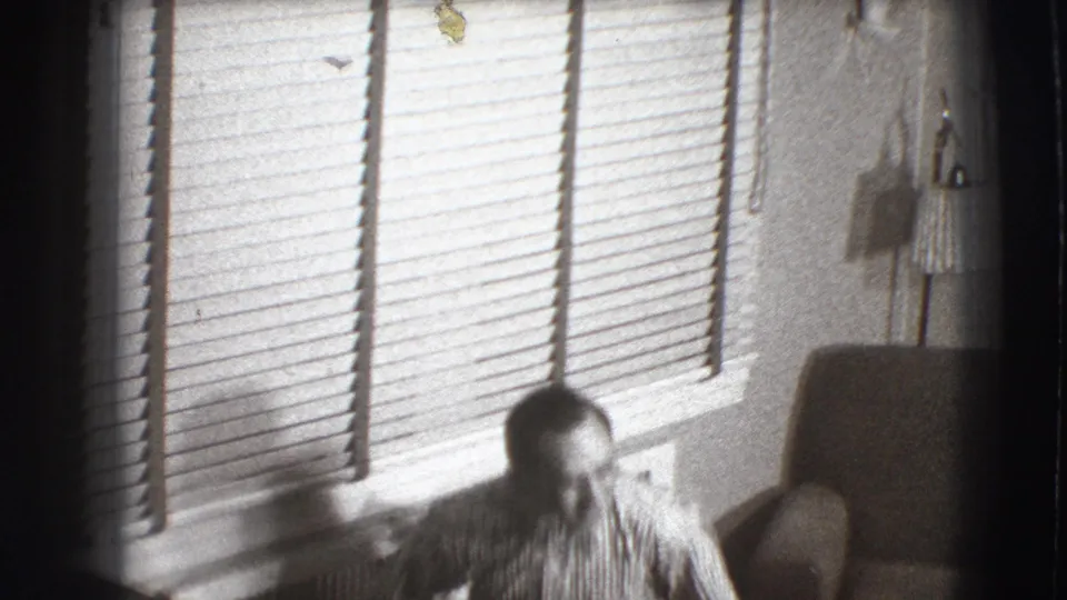 A man in a striped shirt sits in a Joliet, Illinois living room, partly low in the frame near bright window blinds.