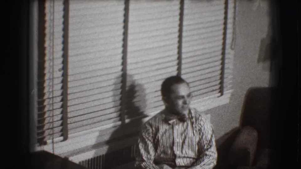 The 1941 home-movie frame shows a seated man in a striped shirt beneath venetian blinds in a private living room.
