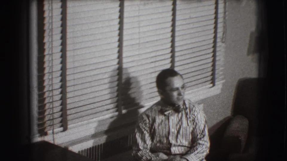 A man sits in profile in the Joliet living room, framed by venetian blinds, wall shadows, and an upholstered chair.
