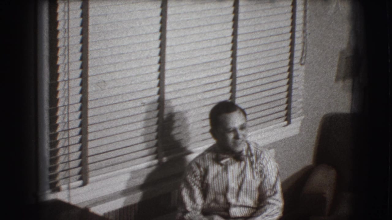 Man in a striped shirt and bow tie sits beside venetian blinds in a dim room.