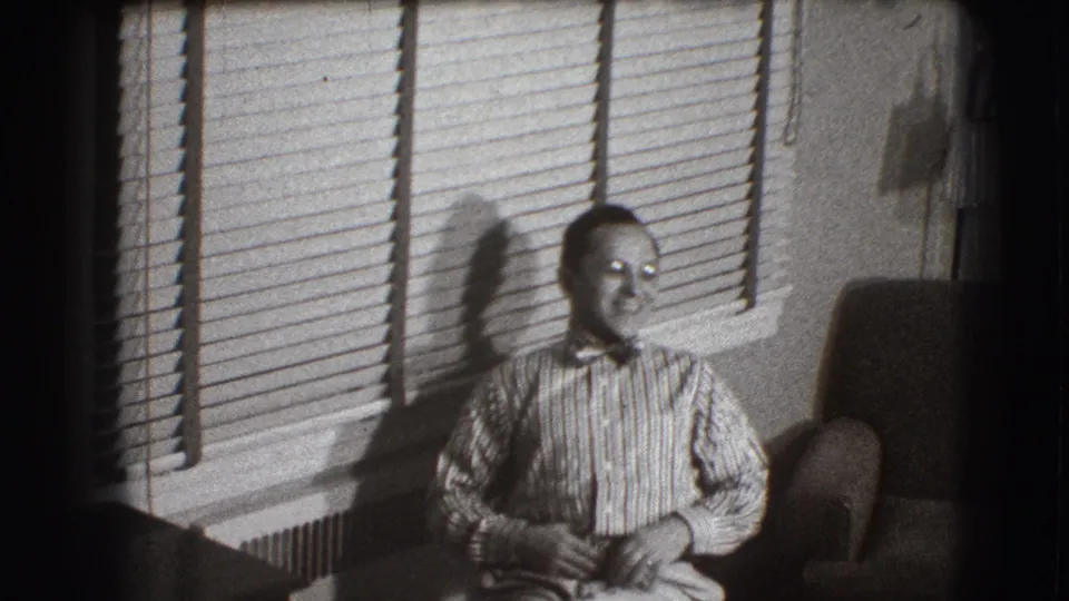 The man sits upright in the Joliet interior, looking off camera as venetian blinds and an armchair frame the scene.