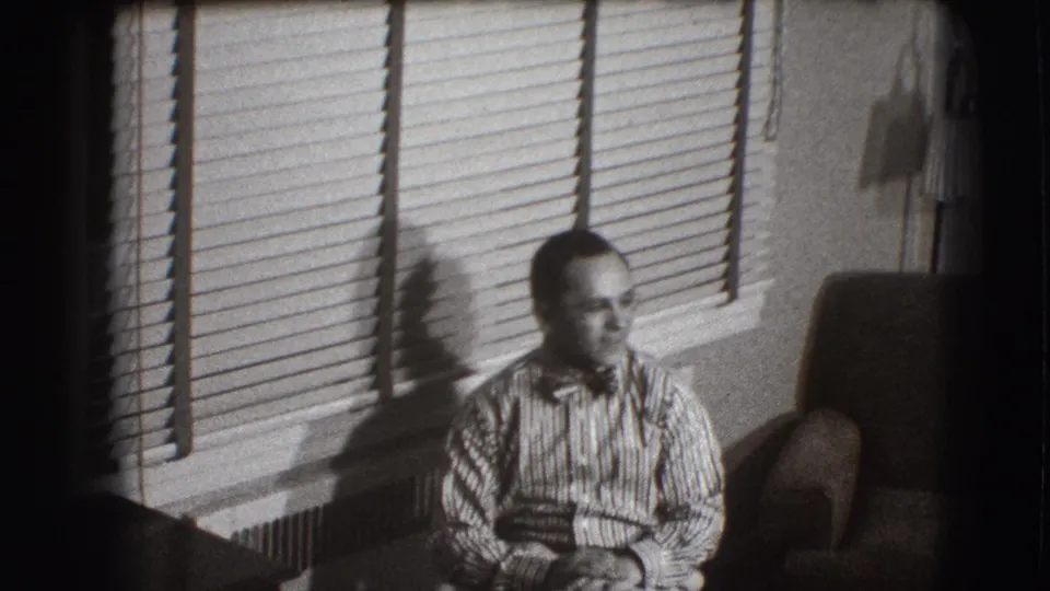 In the 1941 home interior, the man sits with hands together as the blinds create strong horizontal lines behind him.