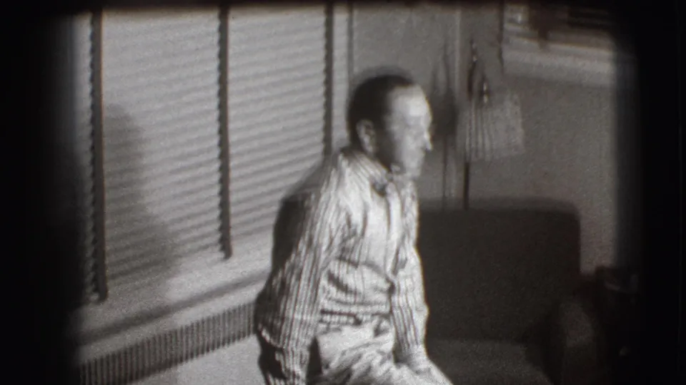 The man leans forward in the dim Joliet room, with venetian blinds, a floor lamp, and furniture visible around him.