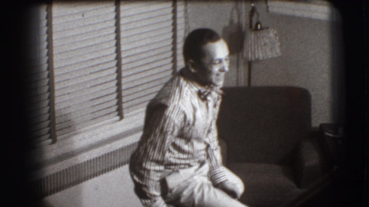 Man with glasses sits sideways near a backward chair in a living room with blinds.
