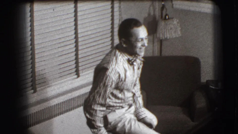 In a 1941 Joliet home interior, a man in glasses and a striped shirt sits awkwardly near a chair beneath venetian blinds.