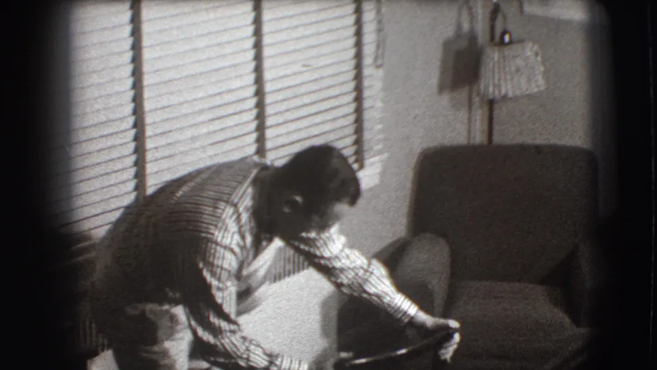 The man leans over the chair back toward his foot inside the Joliet living room, with blinds and an armchair behind him.