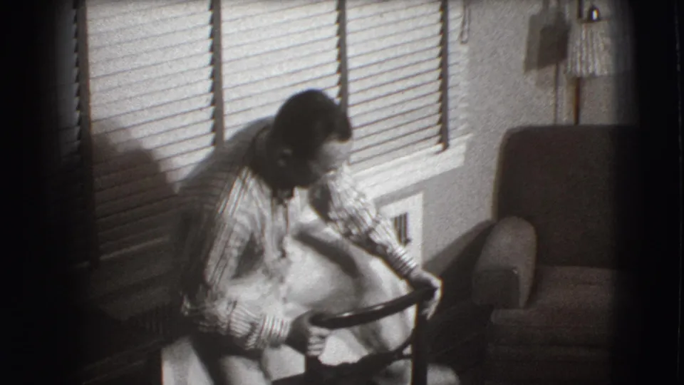 The man remains bent over the backward chair, focused on his foot, in a black-and-white 1941 home-movie interior.