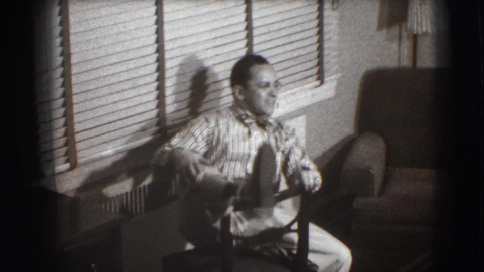 The man sits backward on the chair with one foot raised, looking toward the camera in the Joliet living room setting.