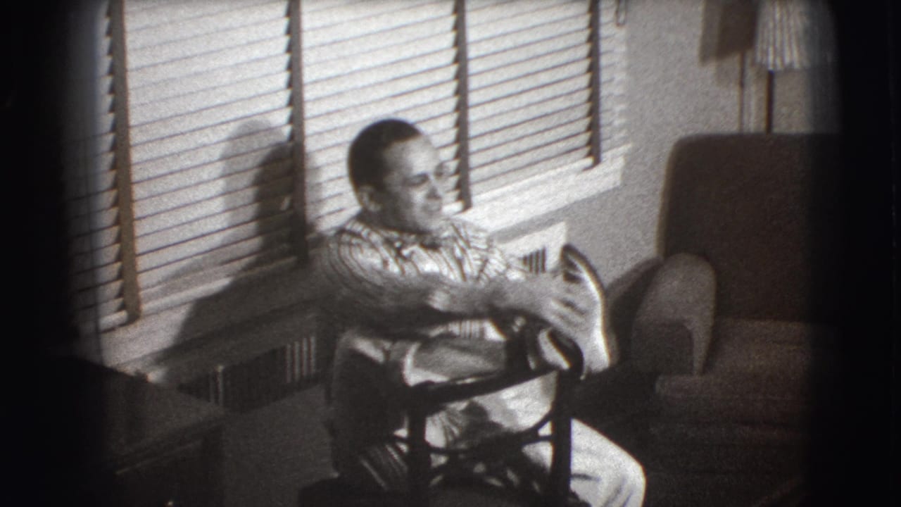 Man with glasses sits by window blinds in a small interior room.