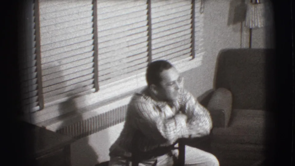 The Joliet patient leans forward in the chair, framed by venetian blinds, radiator, and a nearby upholstered armchair.