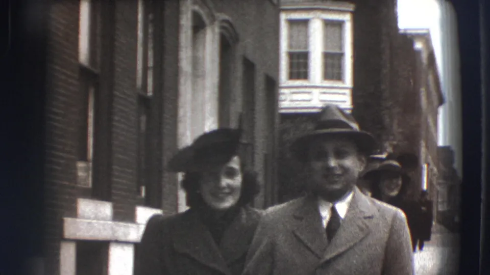 In 1941 Joliet, Illinois, a couple in coats and hats walks toward the camera between brick buildings and a bay window.