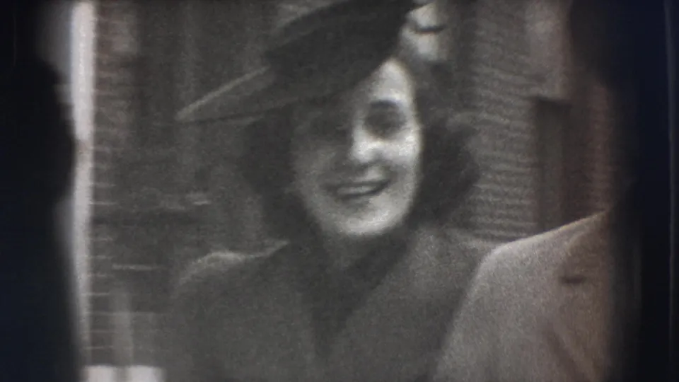 A close 8mm frame shows the smiling woman in a dark angled hat and coat, with the man's shoulder partly visible beside her.