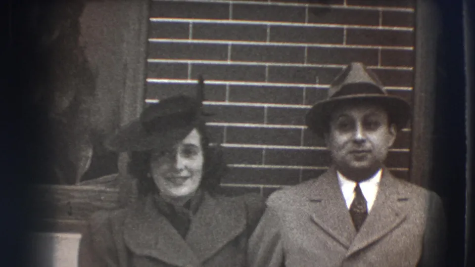 The couple appears to pause near a brick wall and doorway in Joliet, with the man in a suit and fedora and the woman in a dark hat.