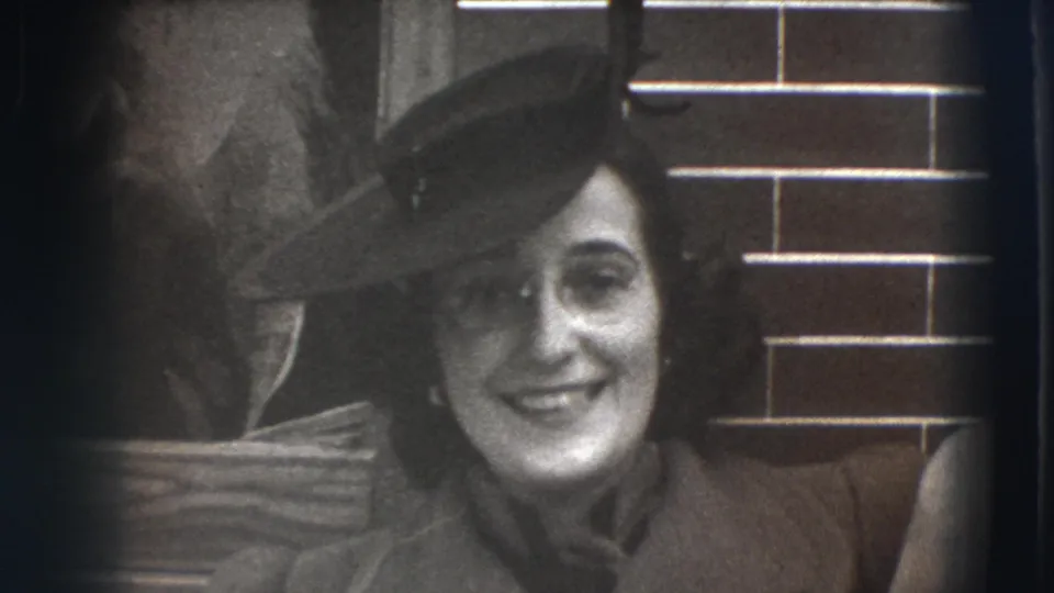 In 1941 Joliet home-movie footage, a woman in a dark hat and coat smiles toward the camera beside a brick entryway.