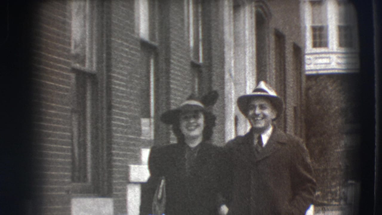 A smiling woman and man in hats stand near a brick building and tall windows.