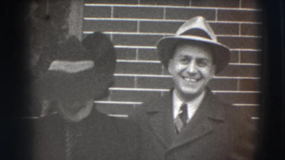 A 1941 Joliet home-movie frame shows the man smiling in front of a brick wall as the woman stands partly obscured nearby.