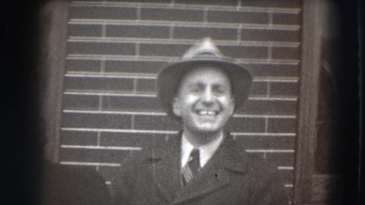 Man in a fedora and suit smiling in front of a brick wall.