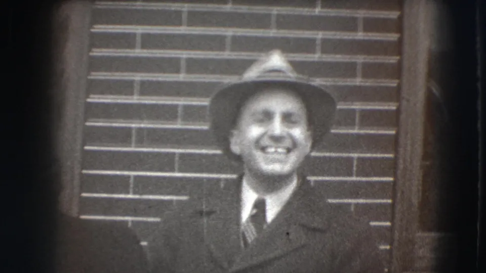 In supplied 1941 Joliet home-movie footage, a suited man in a fedora smiles toward the camera beside a brick wall.