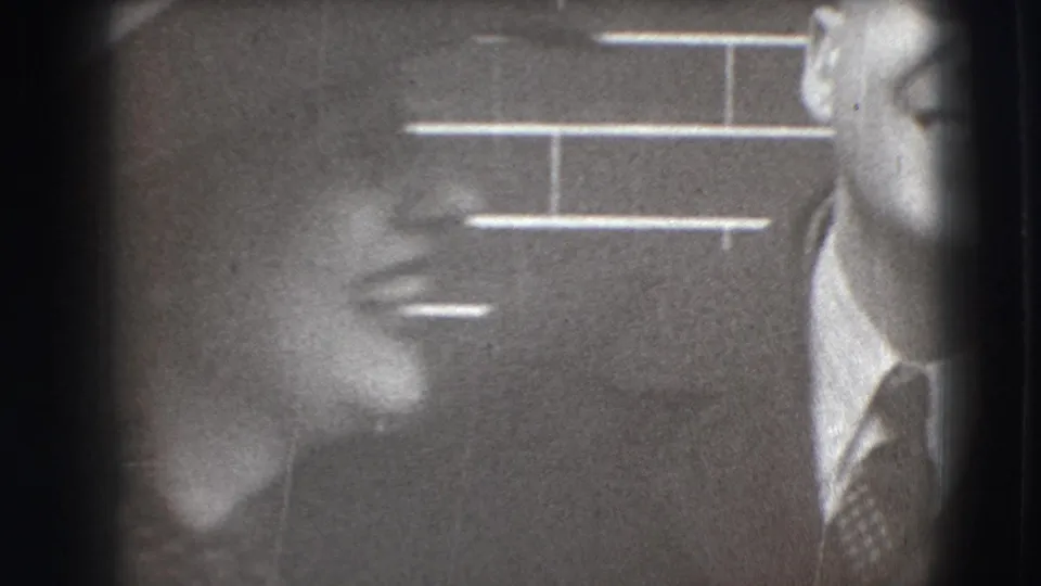 A woman in profile smiles toward a suited man at the edge of the frame, with brickwork visible behind them.