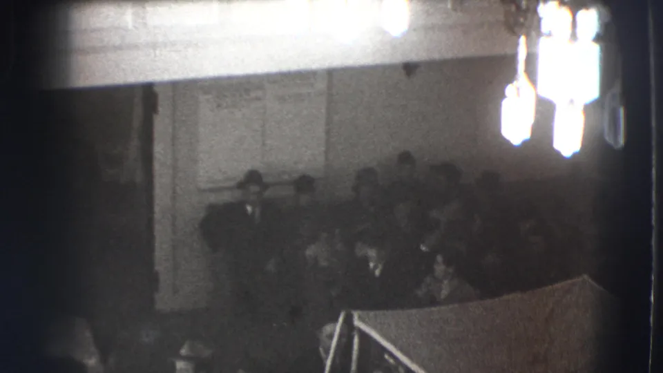 Black-and-white 1941 Joliet frame showing men seated in a dim interior hall beneath bright overhead fixtures.