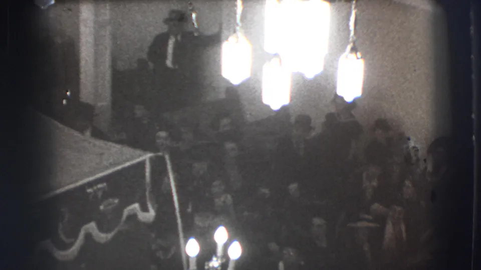 Black-and-white 1941 Joliet frame with a standing speaker addressing seated men in a dim meeting hall.