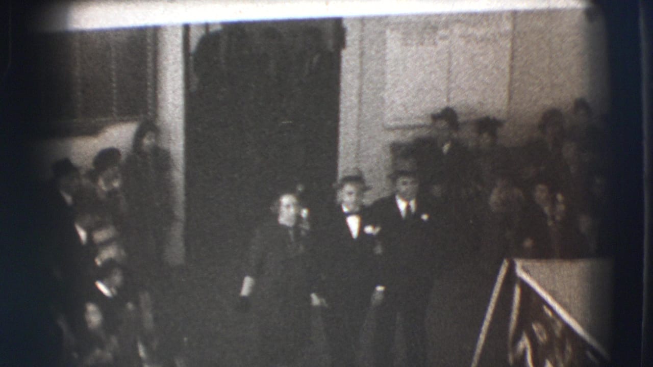 Formally dressed adults stand inside a dim interior near a railing and a large casket-like object.