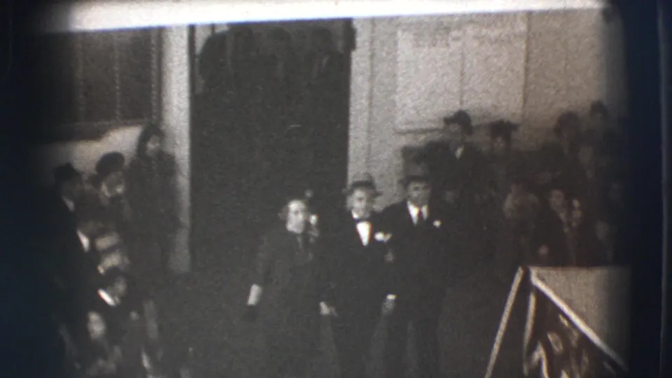 In a 1941 Joliet home movie frame, adults in suits, hats, and dark formal clothing gather inside a dim room near a rail and casket-like form.
