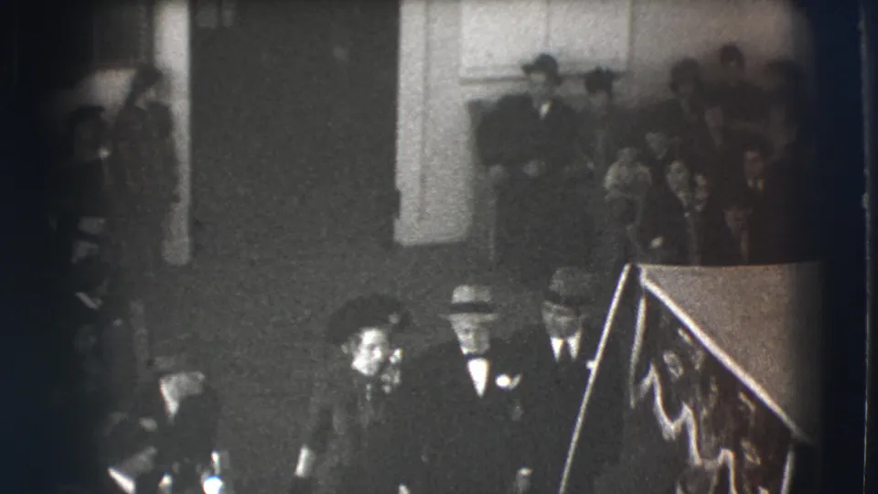 In this Joliet, Illinois 1941 home-movie frame, formally dressed adults pass beneath the camera in a dim interior with onlookers behind them.