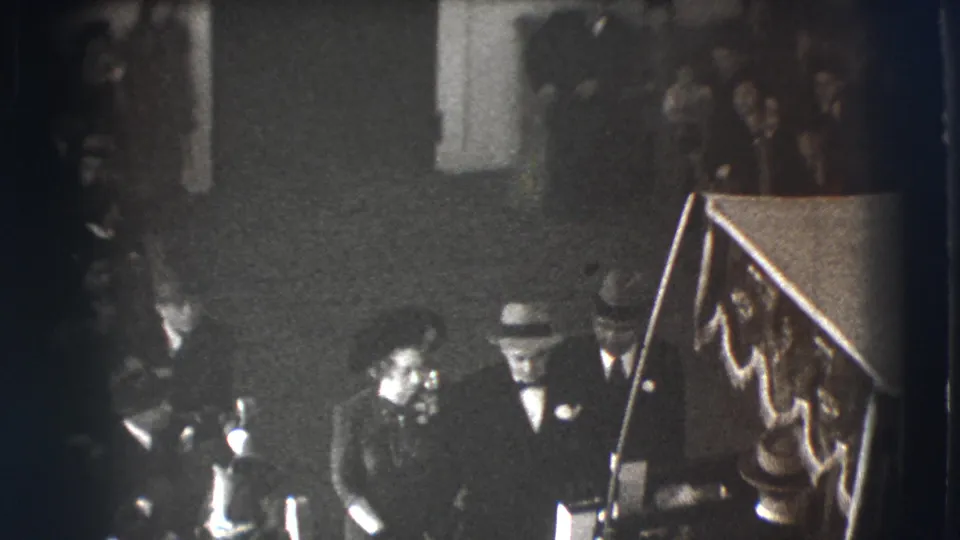 A late frame from the 1941 Joliet clip shows formal attendees beside a rail, with a dim crowd and possible casket area in the foreground.