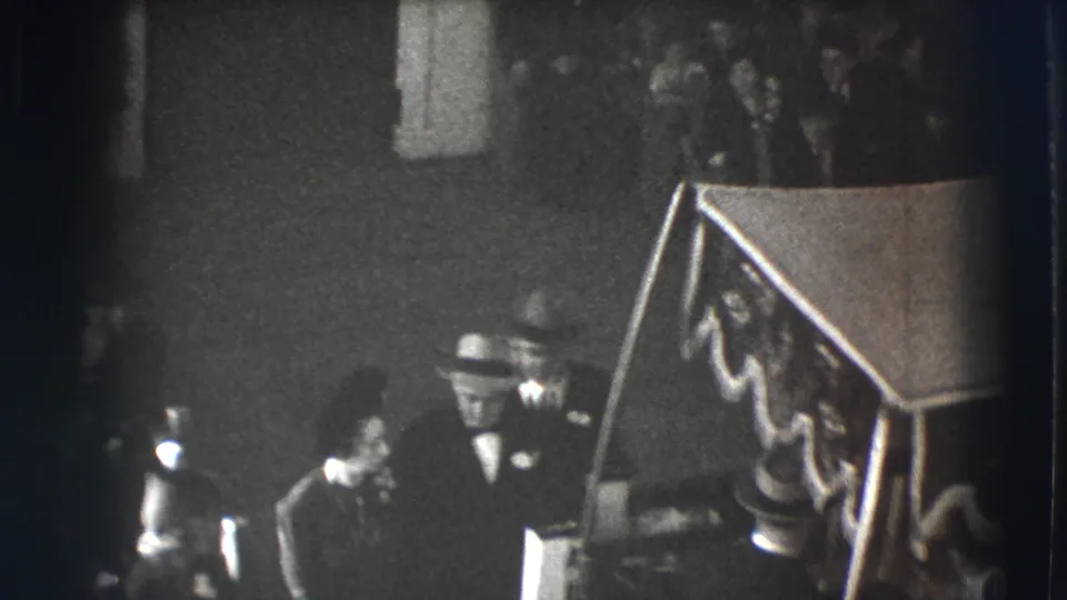 1941 Joliet home-movie frame showing formally dressed men in hats near a decorated stair or podium inside a dim interior.