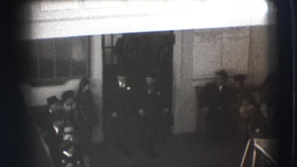 1941 Joliet frame with guests gathered by a doorway and paneled wall, viewed from an elevated home-movie angle.