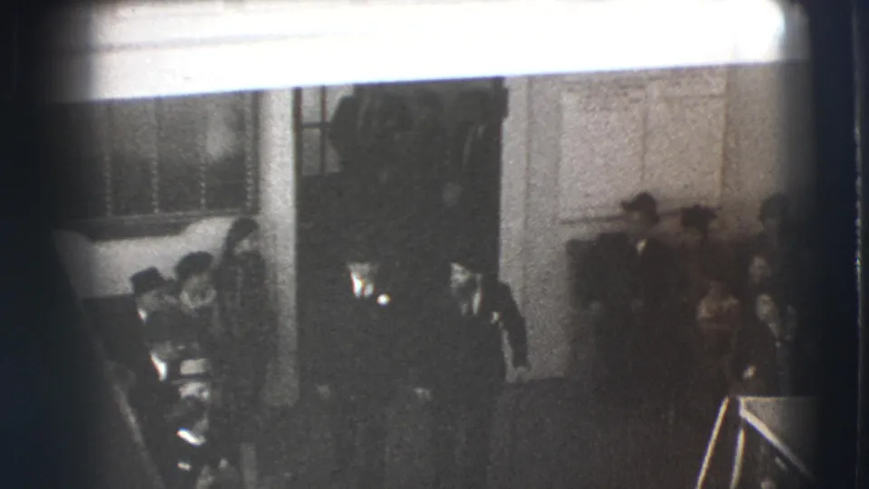 1941 Joliet home-movie frame showing men in suits and hats moving past seated guests inside a formal room.