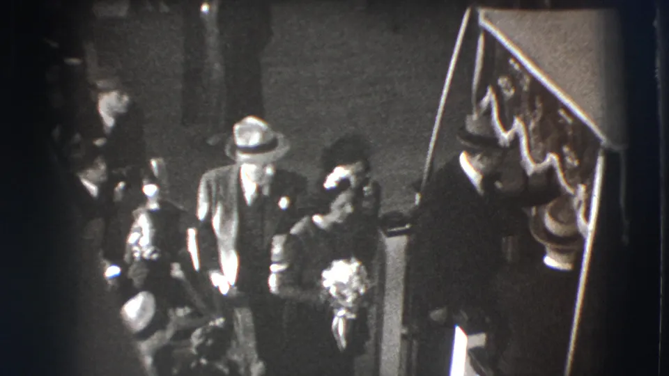 In the Joliet funeral scene, a formally dressed couple stands beside the draped casket while mourners cluster nearby.