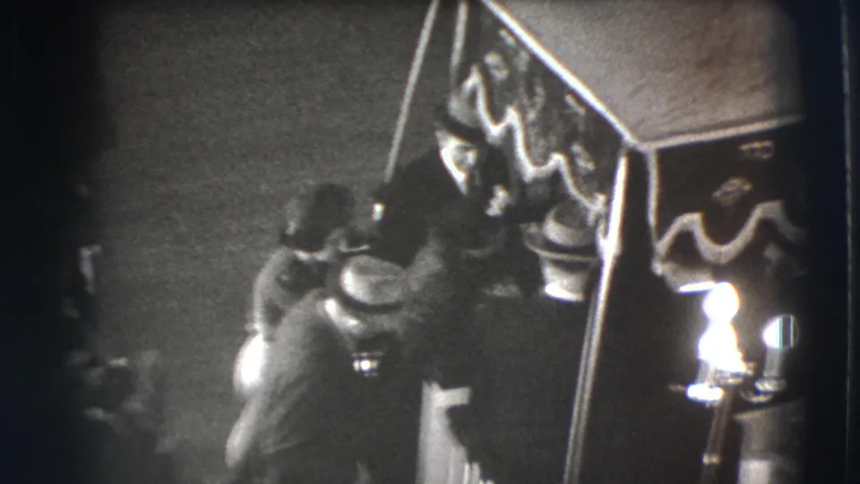 1941 Joliet 8mm frame showing a suited man seated under a draped canopy while another man approaches in a dim indoor gathering.