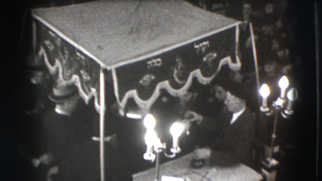 Men in dark suits sit beside a decorated canopy with Hebrew lettering and bright candle flames.