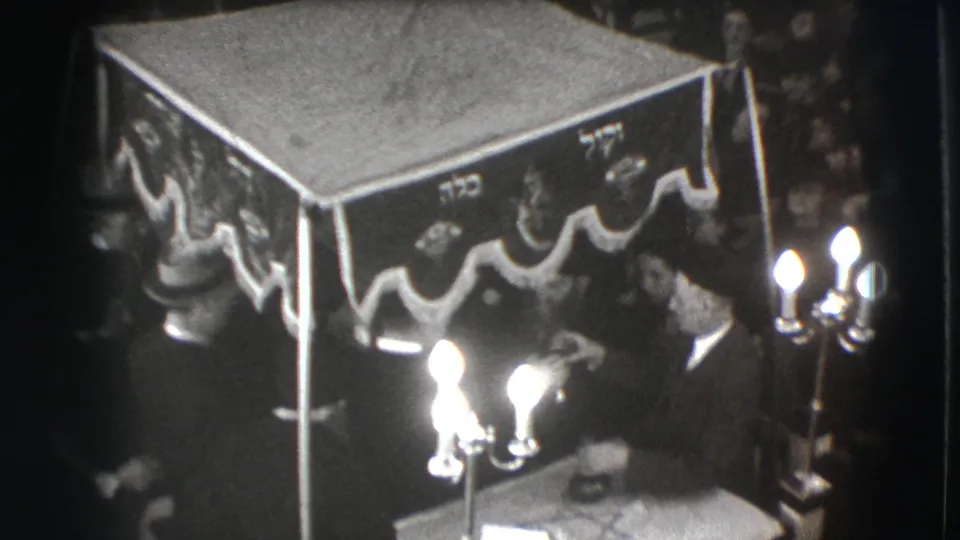 1941 Joliet home-movie frame showing men seated near a Hebrew-inscribed canopy, with candles glowing in the foreground.