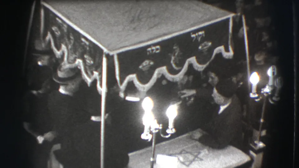 1941 Joliet frame showing a suited man leaning near candles and a Hebrew-inscribed canopy during a formal gathering.
