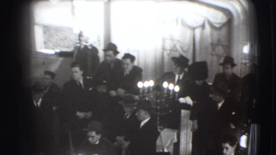 In Joliet, Illinois, 1941, suited men gather in a private interior around lit candles during a Jewish religious observance.