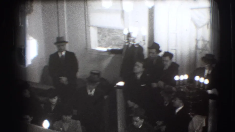 A 1941 Joliet home-movie frame shows suited men in hats gathered near candles, drapery, and Jewish symbolic decoration.