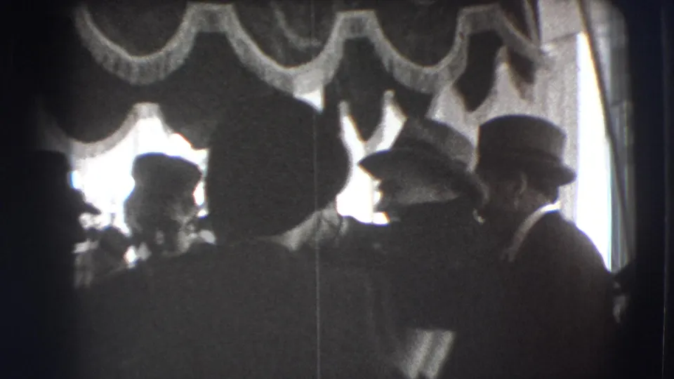 Black-and-white 8mm frame from Joliet showing a crowded ceremonial room with hatted guests and scalloped canopy decoration.