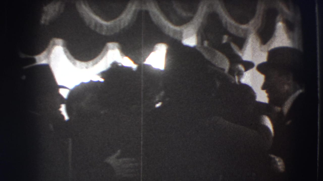 1941 Joliet home-movie frame showing adults in formal dress gathered indoors beneath heavy drapery and bright window light.