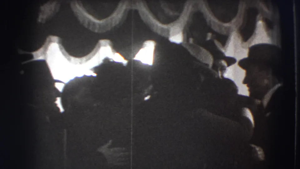 1941 Joliet home-movie frame showing adults in formal dress gathered indoors beneath heavy drapery and bright window light.
