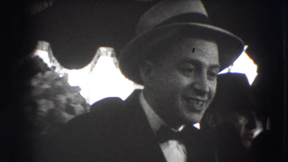 Smiling man in a fedora and bow tie at a 1941 Joliet formal gathering, with other guests and drapery behind him.