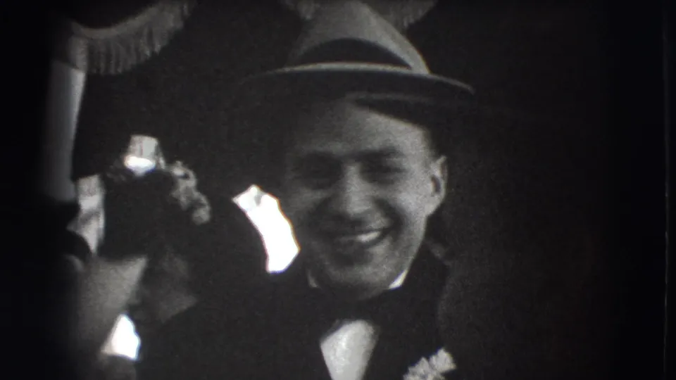 Smiling fedora-wearing guest at a 1941 Joliet formal indoor gathering, framed by nearby adults and bright window light.