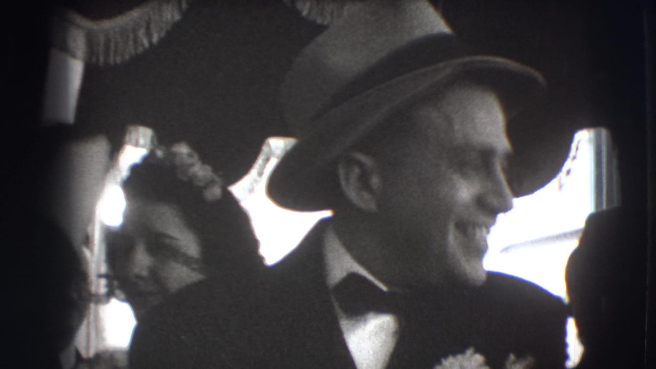 Smiling man in a suit and fedora beside women in floral hats indoors.