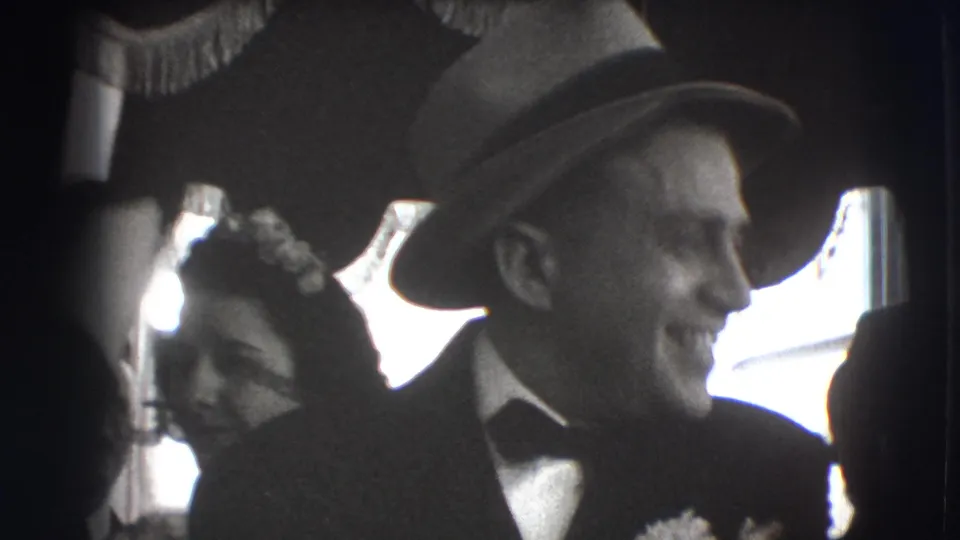 In 1941 Joliet home-movie footage, a smiling man in a suit and fedora stands close to women in dress hats during an indoor gathering.