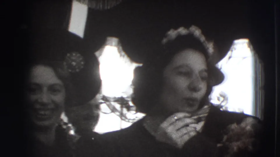 Women in formal clothing and floral hats appear in a dim interior, with a bright window behind them in the 1941 Joliet gathering footage.